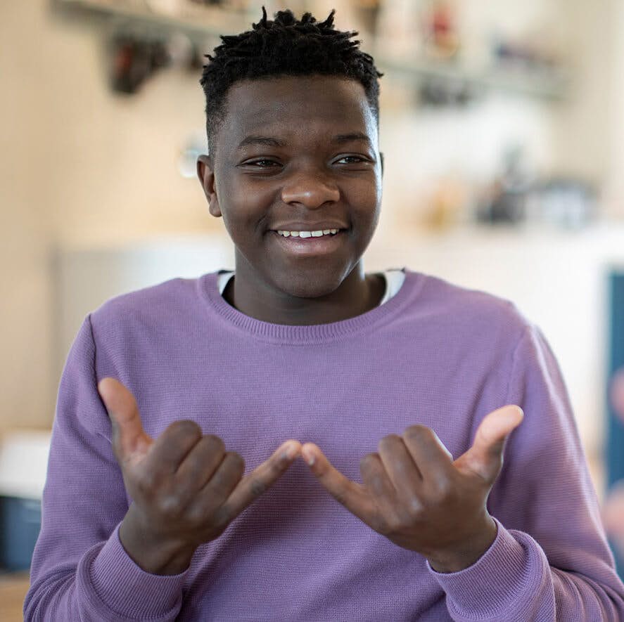 A young African American man dressed in a bright purple sweater smiling while speaking sign language.